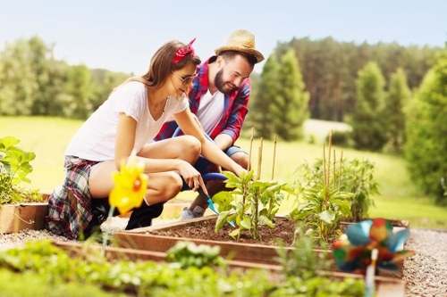 The image depicts a woman and a man working together in a well-maintained garden, situated in an outdoor space with a lush green lawn, tall trees, and a clear blue sky in the background. The woman, wearing a white top and plaid skirt, is planting or tending to young plants in a raised garden bed made of wooden planks, with soil and emerging greenery visible. The man, dressed in a plaid shirt and straw hat, is assisting her, also focused on the garden. Nearby, there are small decorative garden ornaments, and the garden bed is bordered by gravel or small stones. The scene is illuminated by bright natural sunlight, indicating a warm, sunny day. This outdoor garden space is part of a landscaped yard suitable for various gardening activities, highlighting the importance of garden design, plant care, and outdoor maintenance, which Landsapers Swiss Cottage offers in the local area near SW11 postcode and Fulham, London.