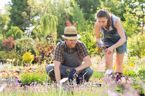 A garden scene featuring two women working together in a well-maintained outdoor space, with one woman wearing a straw hat, a checked shirt, and gardening gloves, crouched down tending to a flower bed filled with various small, colorful flowering plants. The other woman, with long dark hair, dressed in a sleeveless top and denim shorts, is standing and leaning over, holding gardening tools as she appears to assist or observe. The garden includes a lush lawn with dense green grass, bordered by flower beds that contain a mix of flowering plants in shades of pink, purple, and yellow. In the background, there are mature trees and a variety of shrubs, contributing to a vibrant and natural environment. The scene is illuminated by bright, natural sunlight, indicating a clear, pleasant day, and the overall setting reflects a typical outdoor garden maintained by professional landscapers such as Landscapers Swiss Cottage, supporting gardening and outdoor maintenance services in the local area near Swiss Cottage, London.