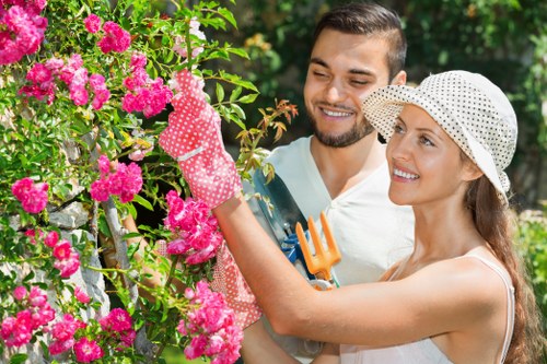 A smiling couple working together in a well-maintained garden in Swiss Cottage, with vibrant pink flowering bushes and lush green foliage in the background. The woman, wearing a white hat with black polka dots and pink gardening gloves, is carefully handling the flowers, while the man, dressed in a light-colored shirt, holds an orange garden fork. The garden features a neat lawn with dense, healthy grass, bordered by flower beds with blooming plants, and a paved pathway partially visible in the background. Natural sunlight illuminates the scene, highlighting the rich green tones of the trees and shrubs, creating a bright, inviting outdoor space that reflects professional gardening and landscaping standards relevant to the Swiss Cottage area. This image exemplifies outdoor garden maintenance, planting, and lawn care services offered by Landscapers Swiss Cottage, situated within the NW3 postal district, beautifully showcasing a vibrant, organized garden environment.
