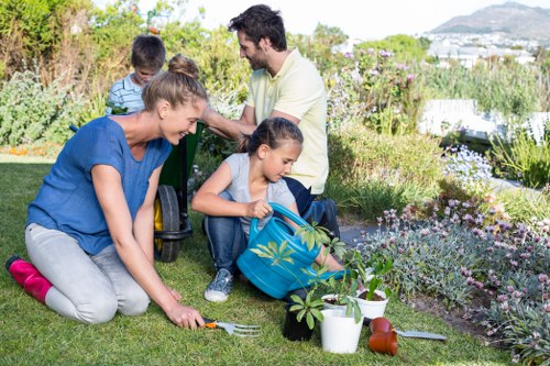 A family gardening together in a private backyard, with two children and two adults engaged in planting and watering activities. The garden features a lush, well-maintained lawn with vibrant green grass, bordered by colorful flower beds containing mixed flowering plants and shrubbery. The children are using gardening tools, such as a small rake, and have potted plants, watering cans, and garden trowels nearby. The adults assist and supervise, with one kneeling on the grass to help a child. In the background, there are mature trees and a view of distant hills or coastline, suggesting a landscaped outdoor space with natural elements. The weather appears sunny and clear, creating a warm and inviting environment suitable for outdoor gardening and family activities, which aligns with professional garden landscaping and maintenance services offered by Landscapers Swiss Cottage in the London area.