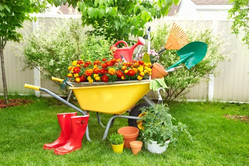 A garden scene in a residential outdoor space during daylight hours, featuring a vibrant yellow wheelbarrow filled with colourful flowering plants, including red and yellow blooms. The wheelbarrow is positioned on a well-maintained grassy lawn, with gardening tools such as a red watering can, a green garden rake, and an orange watering can resting inside and around it. A pair of bright red rubber boots is placed near the wheelbarrow on the grass. To the right, there is a potted plant growing in white containers, and several small terracotta pots are scattered nearby. In the background, a white fence surrounds the garden, with lush green trees and shrubs providing natural shade and privacy. The scene appears to be in a sunny, mild weather condition, with fresh greenery indicative of ongoing gardening or outdoor maintenance work, as might be provided by a professional landscape or gardening service in Swiss Cottage.