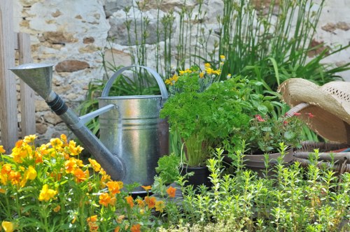 The image depicts a quaint garden scene with a backdrop of a textured stone wall. In the foreground, there is a dense patch of vibrant yellow and orange flowers, likely marigolds, adding bright splashes of colour. Behind these flowers, a well-maintained lawn area with lush, green grass occupies part of the garden space, bordered by a variety of shrubs and taller plants. Notably, a classic metal watering can with a curved handle and extended spout is situated among the foliage, suggesting ongoing garden maintenance. To the right, a woven straw hat rests next to potted plants that include a mix of leafy herbs or flowers, providing a natural, rustic feel. The garden layout combines neatly trimmed plants and a mature, textured stone wall, creating a charming outdoor area compatible with professional garden and landscaping services, such as those provided by Landscapers Swiss Cottage. The scene is bathed in natural daylight, indicating a clear, probably mild weather condition, further enhancing the inviting and well-kept appearance of the garden environment in an outdoor space within the vicinity of Swiss Cottage, NW3 postcode area.