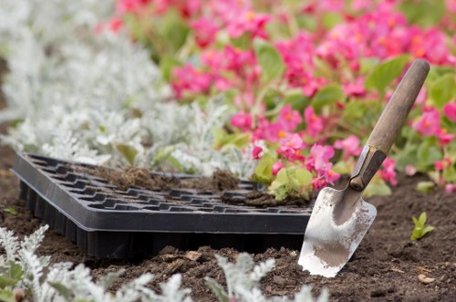 An outdoor garden scene featuring a section of freshly turned soil with a small gardening trowel embedded in the earth, and a black seed tray resting on the soil surface. Surrounding the area are vibrant flower beds with pink and white flowering plants, creating a colourful background. The garden layout includes a neatly maintained lawn area with lush, green grass visible in the foreground, and a variety of shrubs and trees in the distance, indicating a landscaped backyard or front garden typical of properties in Swiss Cottage. The weather appears bright and clear, with natural sunlight illuminating the scene, highlighting the rich textures of the soil, foliage, and garden tools. This image reflects a well-organised, appealing outdoor space suitable for planting or garden maintenance, illustrating the kind of work offered by landscapers in the Swiss Cottage area.