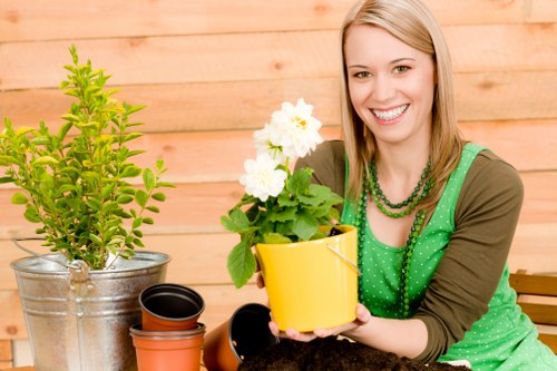 A woman with blonde hair and a bright smile is seated outdoors in a garden setting, holding a yellow ceramic pot containing a green leafy plant with white flowers. Behind her, there is a wooden garden fence and various gardening items including a silver metal bucket with a lush green shrub, empty terracotta pots, and a small black pot. The garden features a well-maintained lawn with dense, green grass in the foreground, bordered by a flower bed or planting area. The environment appears well-lit with natural daylight, suggesting a mild weather day suitable for outdoor gardening activities. The scene reflects a tidy and vibrant outdoor space, typical of residential gardens in the area serviced by Landscapers Swiss Cottage, emphasizing garden planting, container gardening, and outdoor plant care as part of their gardening services.