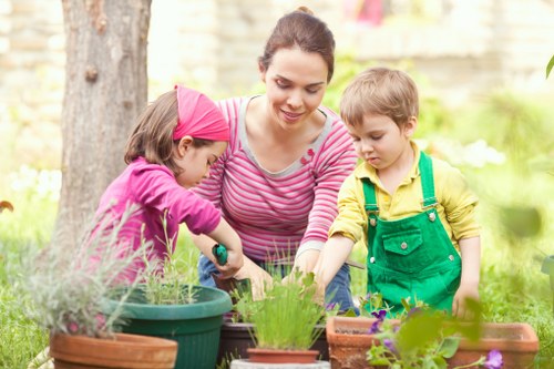 A woman and two children are engaged in gardening activities in a garden setting, with the woman smiling as she supervises their work. The children, a girl dressed in pink and a boy in yellow with green overalls, are planting or tending to potted plants, which include herbs and flowering plants. The garden features a lush green lawn, with various potted plants arranged on a wooden surface or garden table. In the background, there are mature trees and a stone wall, indicating an outdoor space with natural light and a calm, sunny atmosphere. The scene exemplifies outdoor family gardening, aligning with landscapersswisscottage.co.uk's gardening services for maintaining and enhancing gardens in the local area, such as the nearby town or postcode region.