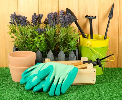 This image displays a neatly arranged gardening setup in an outdoor space, likely within a garden in Swiss Cottage. In the foreground, there is a patch of well-maintained artificial grass, providing a vibrant green base. Behind this, several lavender-colored flowering plants with tall, slender stems are planted in black containers, adding natural colour and vertical interest to the scene. To the left, two empty terracotta pots of different sizes are stacked and ready for planting. Central to the image is a wooden storage box, holding a pair of bright turquoise gardening gloves and a small hand rake, suggesting active gardening work. To the right, a yellow plastic bucket contains different gardening tools, including a trowel and a hand fork, with the handles visible at the top. This outdoor workspace is set against a wooden fencing backdrop, indicating a private garden area suitable for landscaping or maintenance by companies like Landscapers Swiss Cottage, which offers comprehensive gardening services in the local area. The lighting suggests a clear, daylight scene perfect for outdoor gardening activities, emphasizing the preparation and care involved in maintaining a healthy, attractive garden environment.